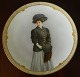Platte i 
porcelæn fra 
Royal 
Copenhagen med 
bemalet portræt 
af kvinde i 
overtøj. 
Platten ...