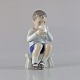 Figur i 
porcelæn med 
motiv af en 
dreng som 
drikker af krus 
og sidder på en 
skammel no ...